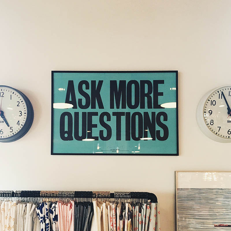 A sign reading "ASK MORE QUESTIONS" in all-caps on a wall between two analog clocks.