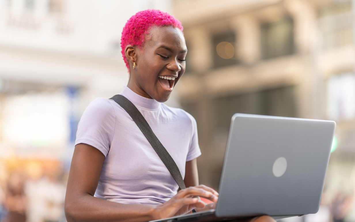 A young woman with vibrant pink hair smiles excitedly while using a laptop. She is sitting outdoors, wearing a light purple t-shirt and a crossbody bag strap over her shoulder. The background is blurred, showing an urban setting with soft lighting and people moving in the distance.