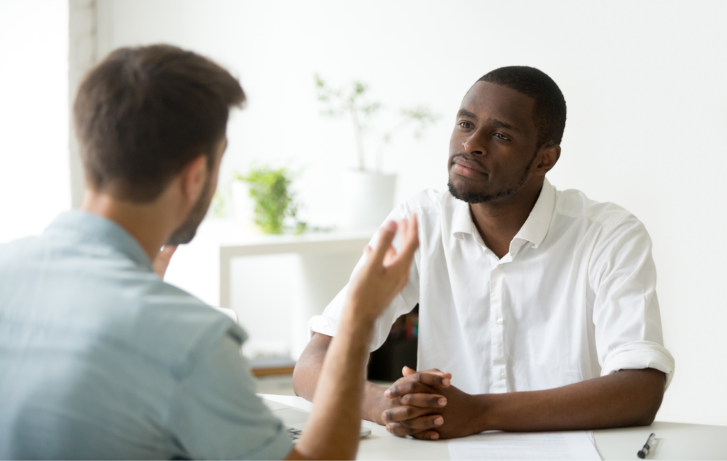 Two men seated at a table engaged in conversation; one listens intently while the other speaks.